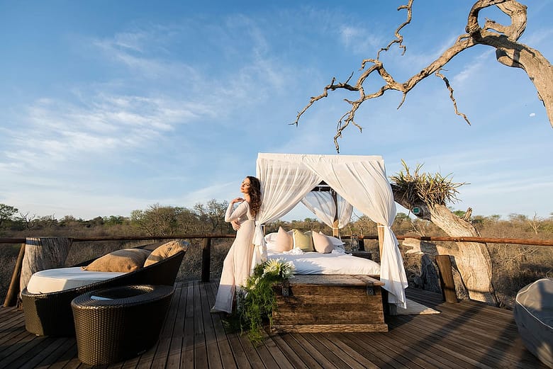 Bride standing next to an out door bed covered in a romatic canopy in the middle of the bushveld on a raised platform